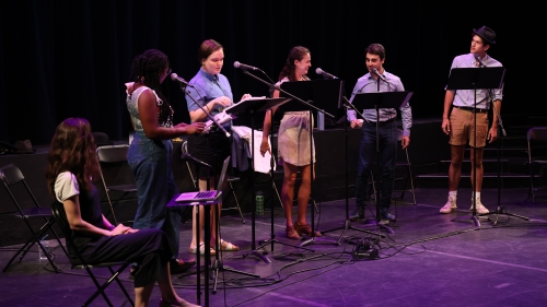 Six students are standing on a stage with stands and scripts.